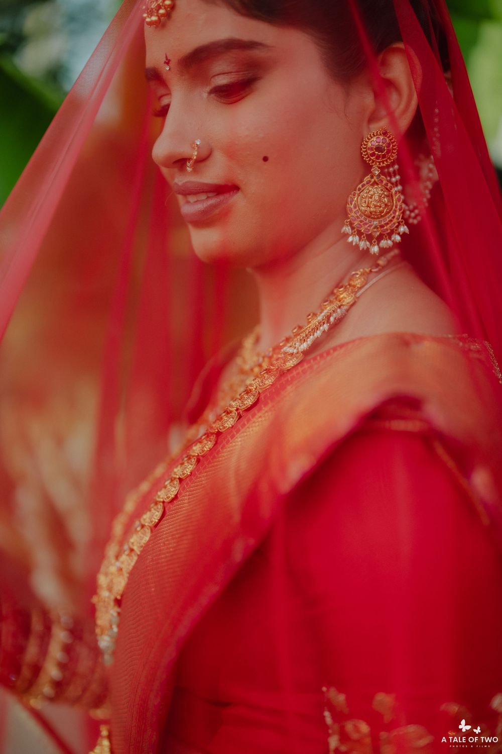 Beautiful Indian bride in red lehenga with gold chandbali earrings and floral necklace under red veil.