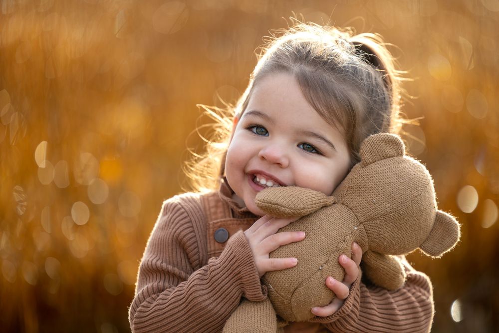 meisje (2) gekleed in bruine kleding, knuffelend met haar teddybeer.