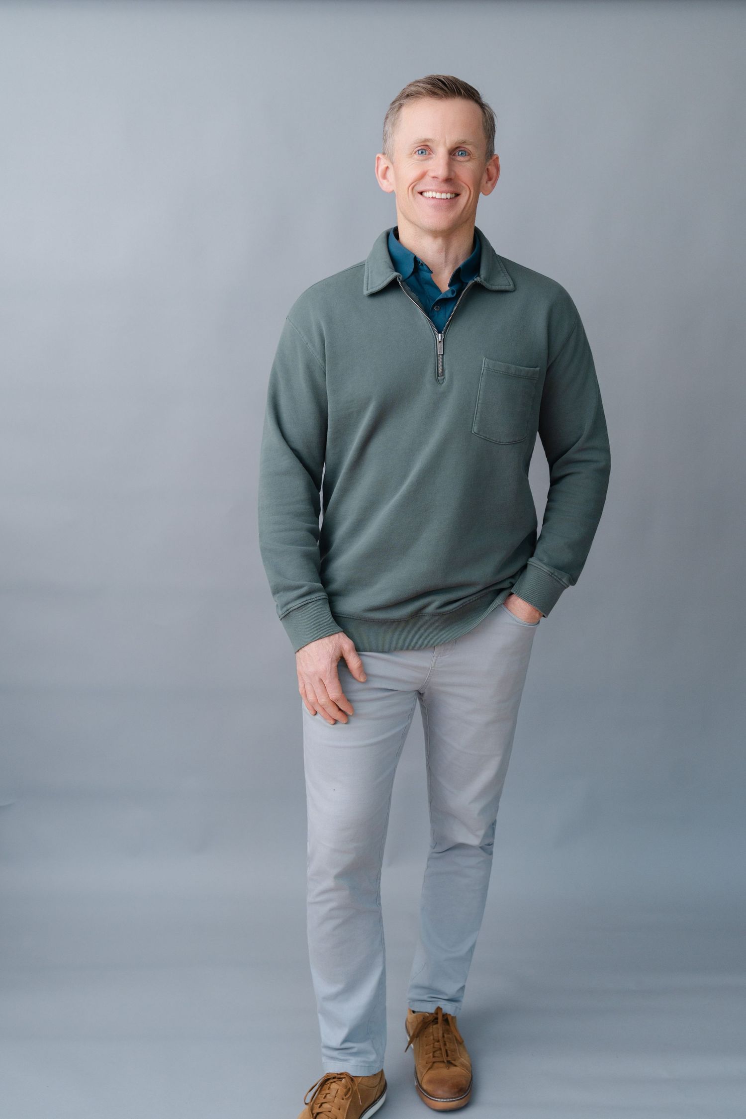 Smiling man in green quarter-zip pullover and light gray pants standing against a gray background.