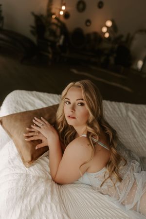Young blonde woman in white lace lingerie reclining on white bed with brown pillow in moody lit room.