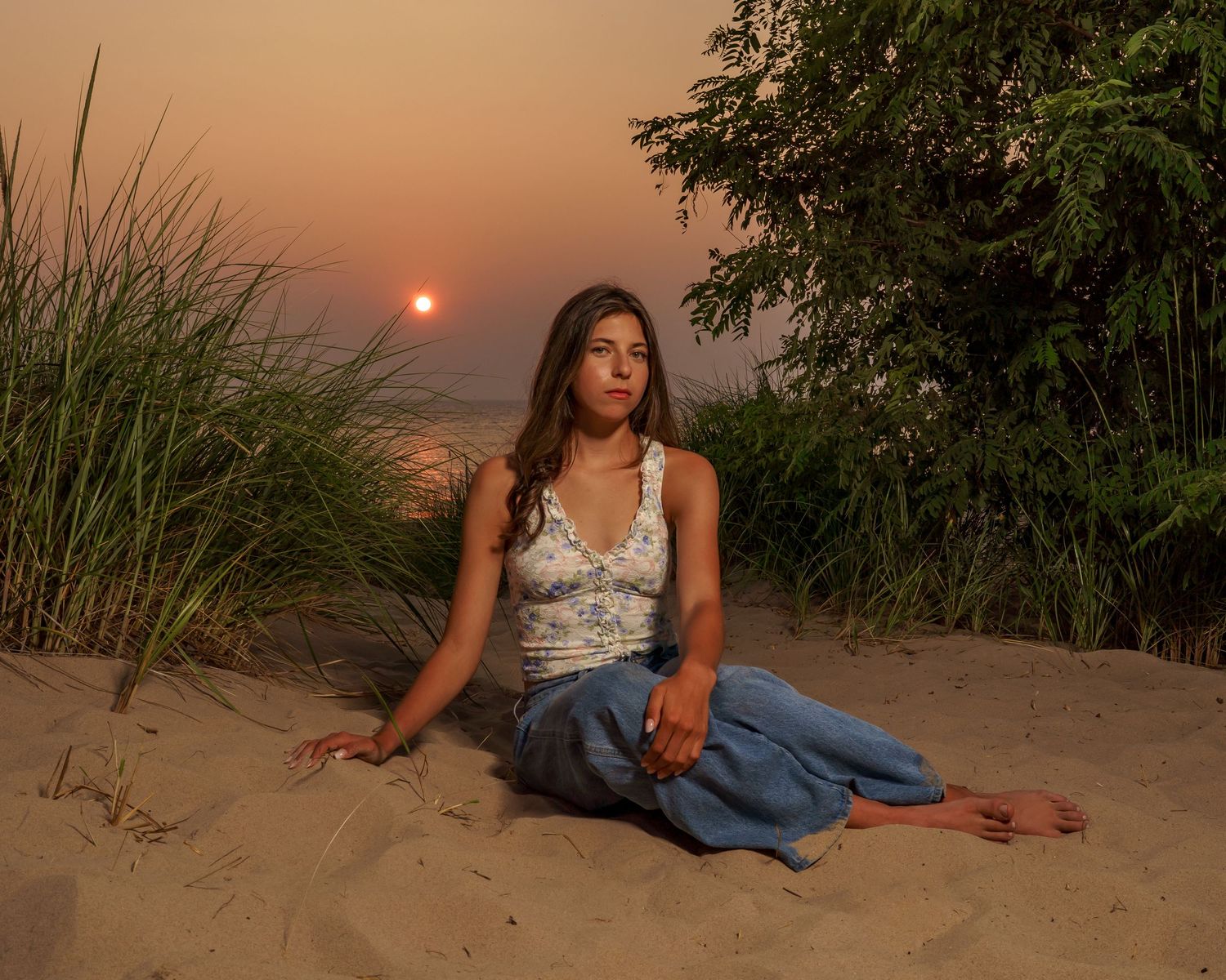 Woman sitting on sandy beach at sunset, wearing blue pants and floral top, with tall grass and trees in background.