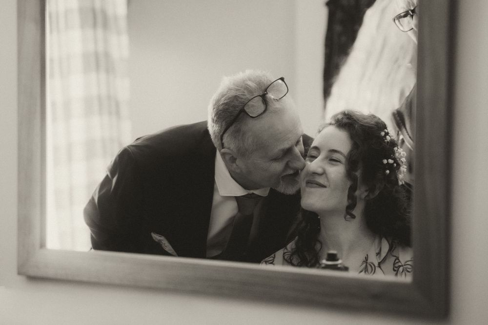 Black and white wedding photo of bride and older man sharing a tender moment, reflected in a mirror.
