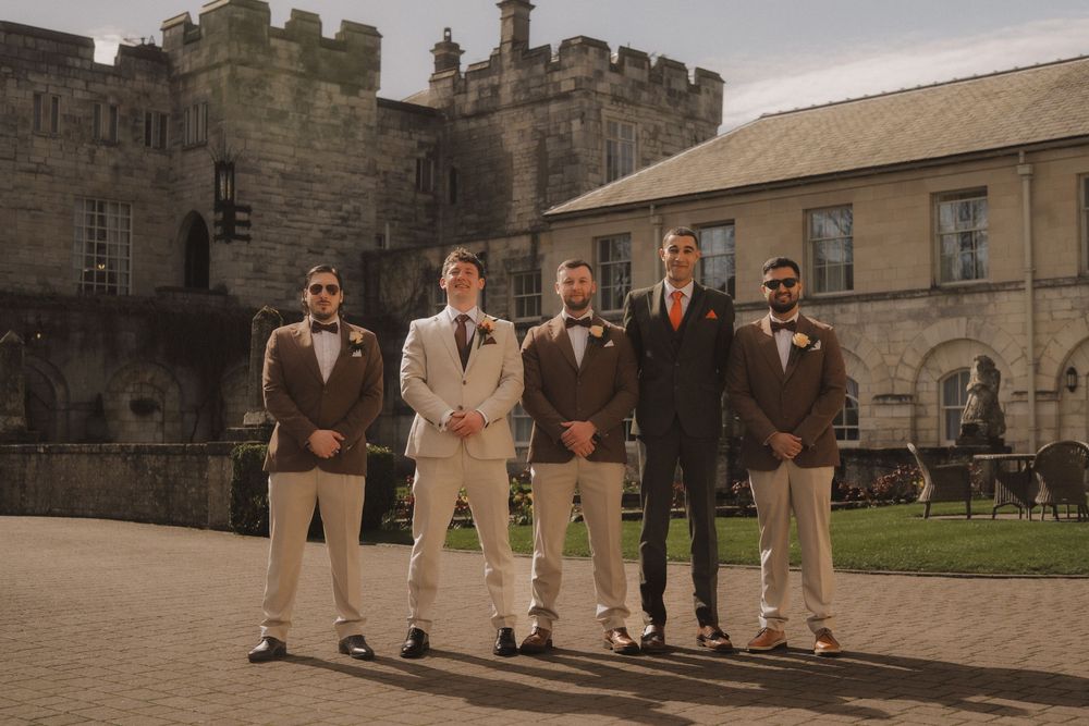 Groom and groomsmen in tan suits and brown bow ties posing outdoors at a castle wedding venue.