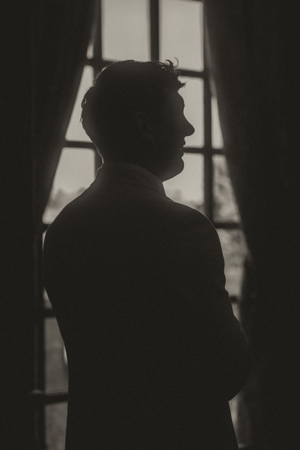 Dramatic black and white silhouette of a man standing near a window with curtains in a dark room.