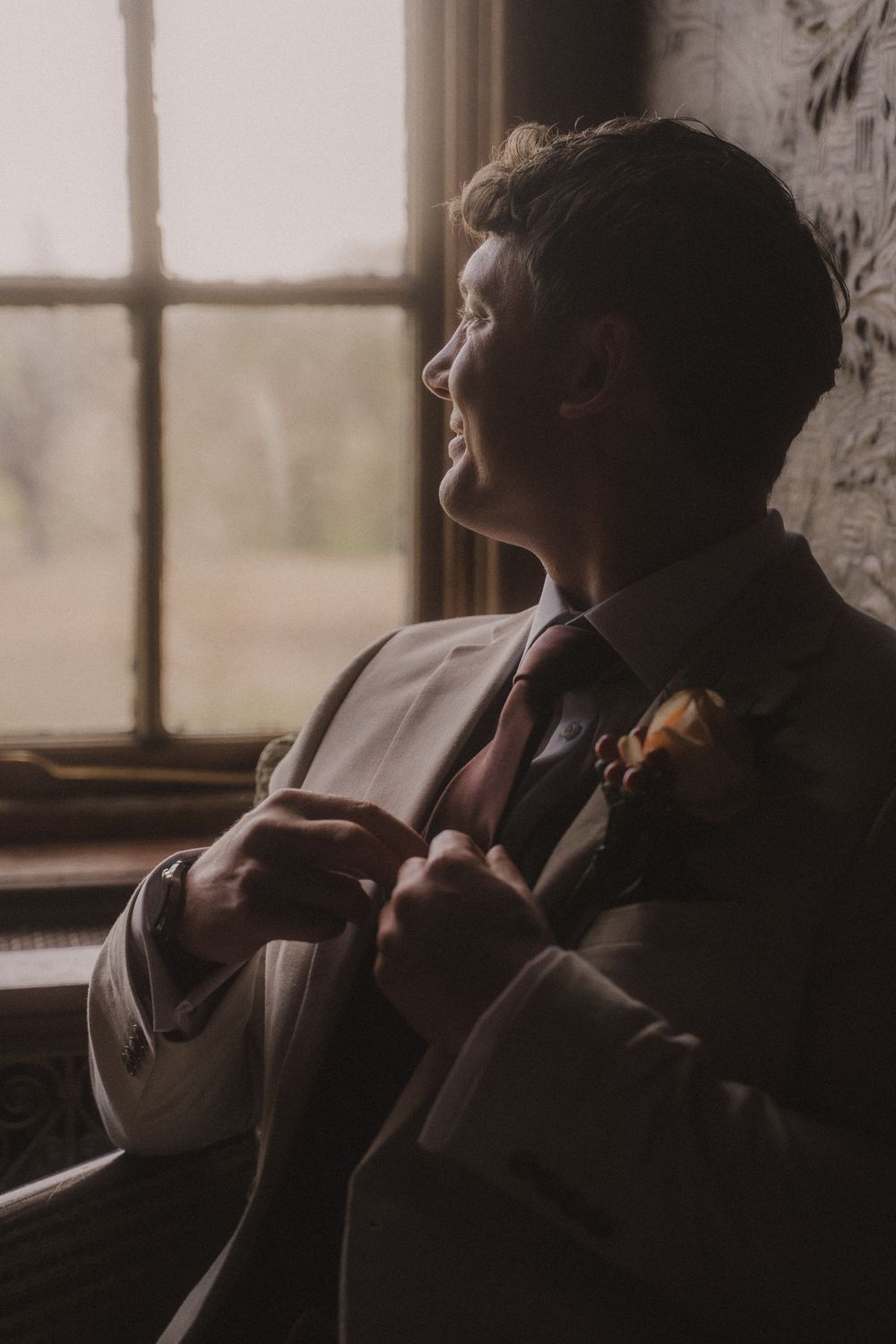 Groom in gray suit adjusting tie by window, wearing boutonniere, soft natural light, elegant wedding portrait.