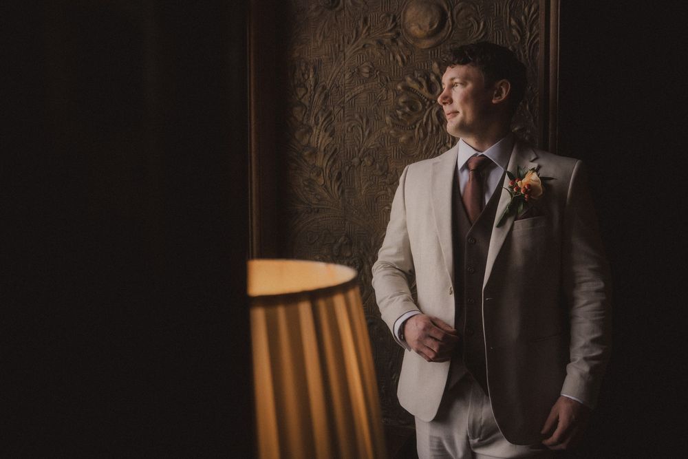 Groom in a cream suit with boutonniere stands by a window in moody, warm lighting against an ornate wall.