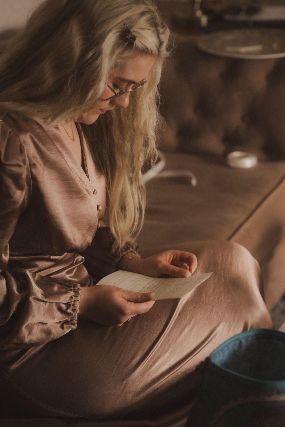 Blonde woman in glasses wearing a mauve dress, sitting cross-legged on a sofa reading a letter indoors.