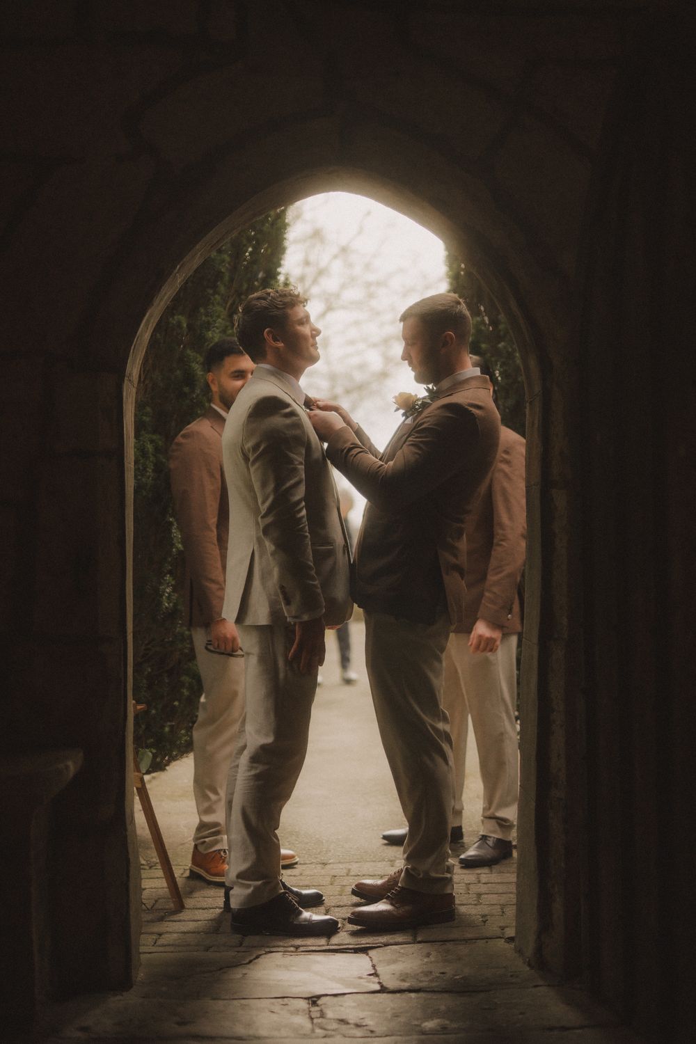 Groomsmen adjusting boutonnieres in a stone archway, wearing brown and grey suits at a wedding.