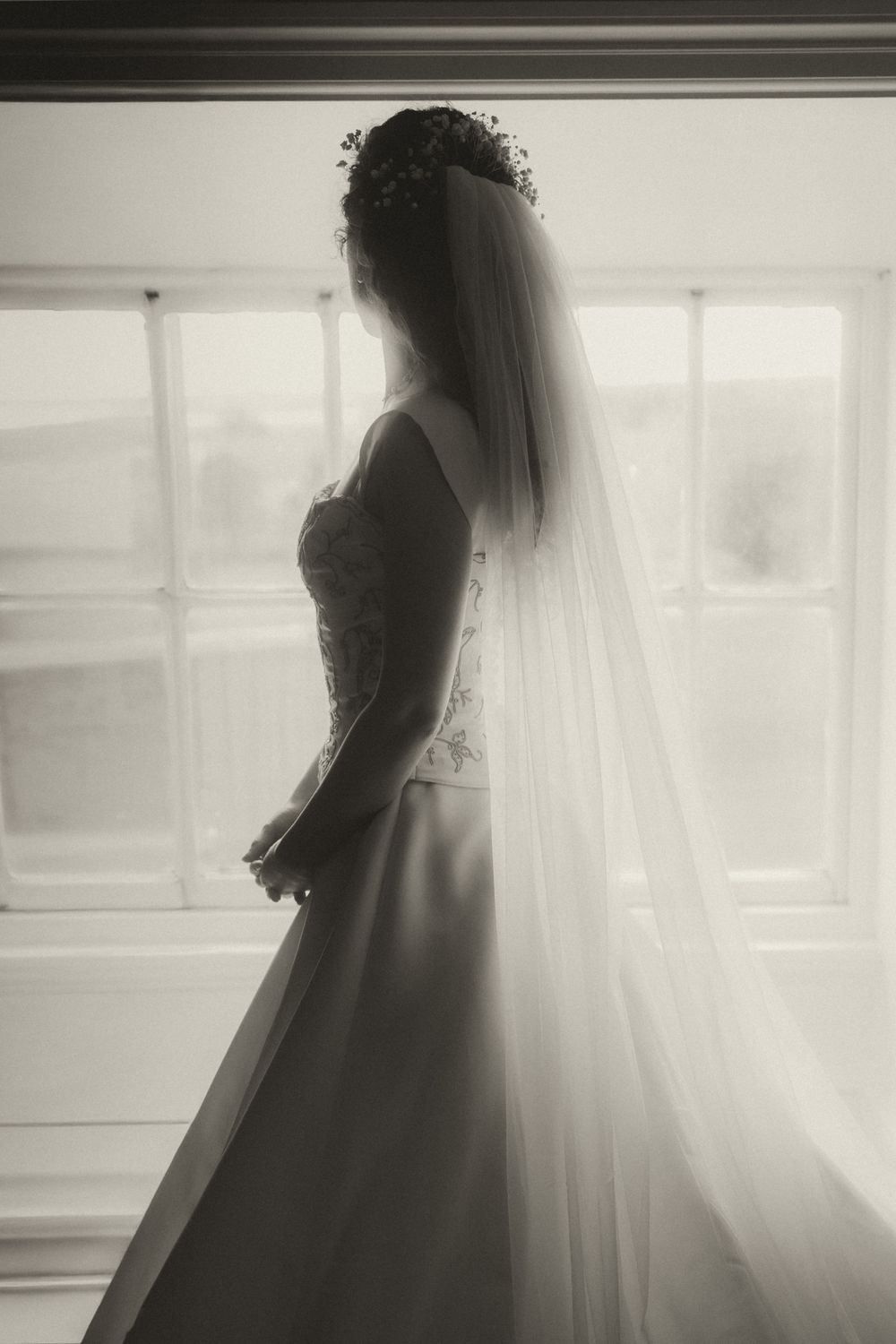 Black and white silhouette of a bride in a wedding gown and veil standing before a window on her wedding day.