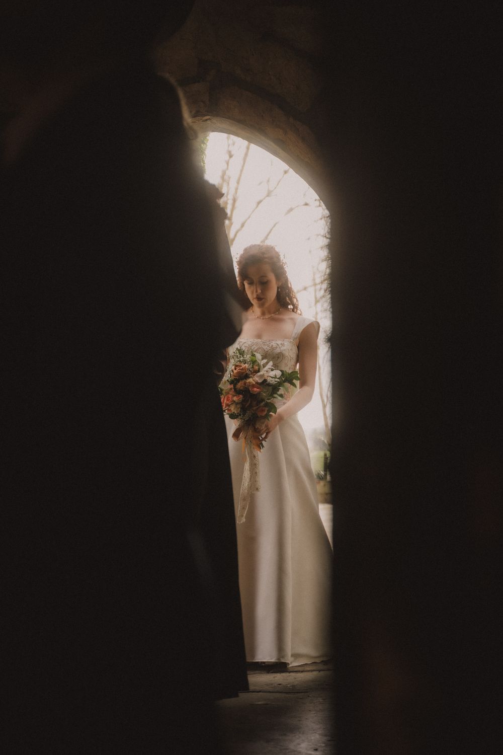 Bride in white gown holding floral bouquet framed by stone archway in dramatic dark and light contrast.