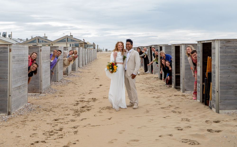 Een gelukkig bruidspaar loopt tussen rijen strandhuisjes door, terwijl juichende gasten op hun trouwdag naar buiten gluren.