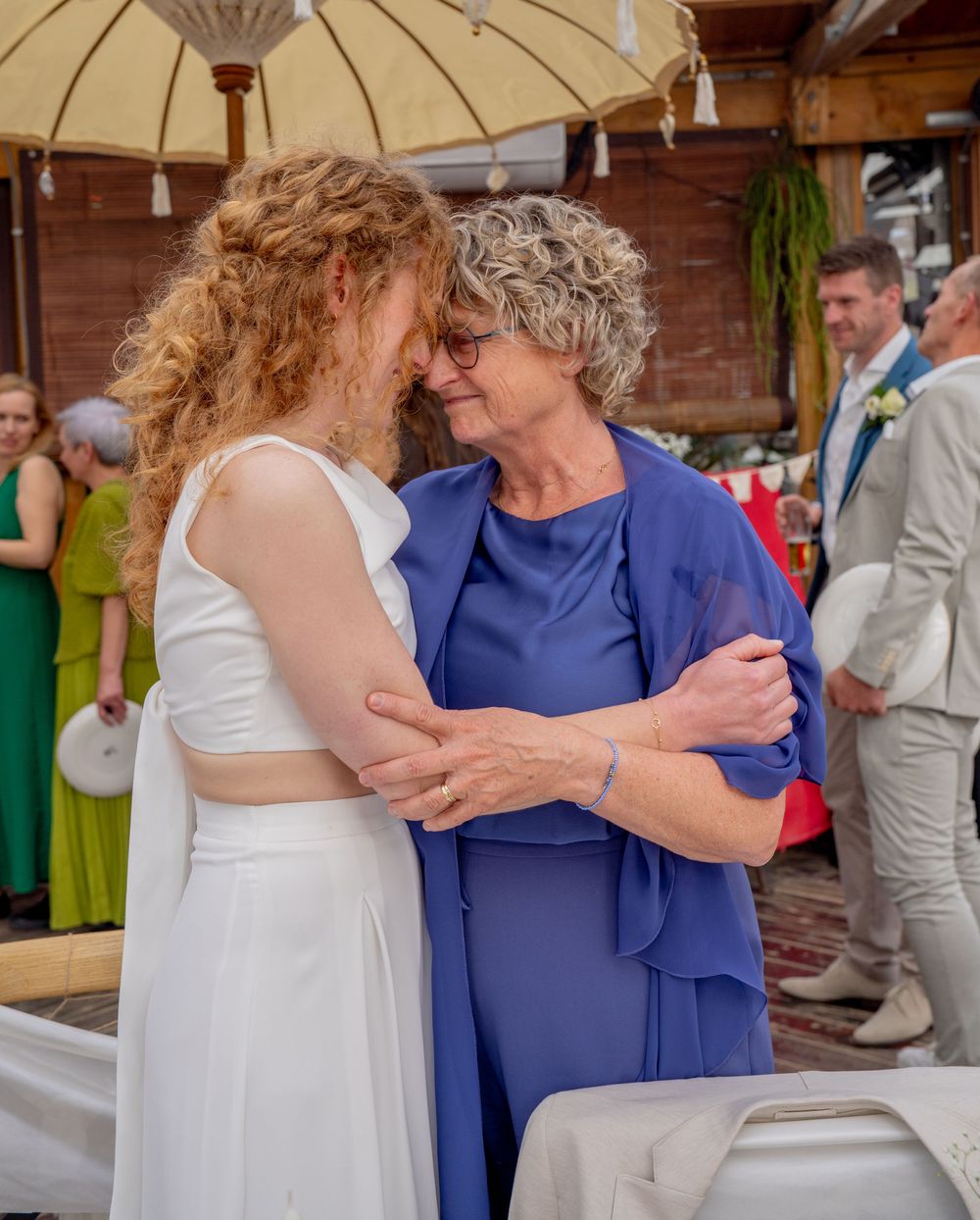 Bruid in witte jurk deelt een tedere omhelzing met oudere vrouw in blauwe outfit tijdens huwelijksreceptie in de buitenlucht.