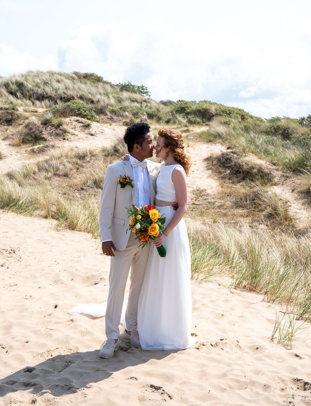 Bruid en bruidegom delen een romantisch moment op de zandstranden, de bruid houdt een levendig geel en oranje boeket vast.