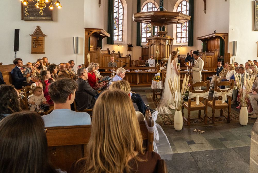 Breedbeeldopname van een volle kerk die het bruidspaar tijdens de huwelijksceremonie gadeslaat.
