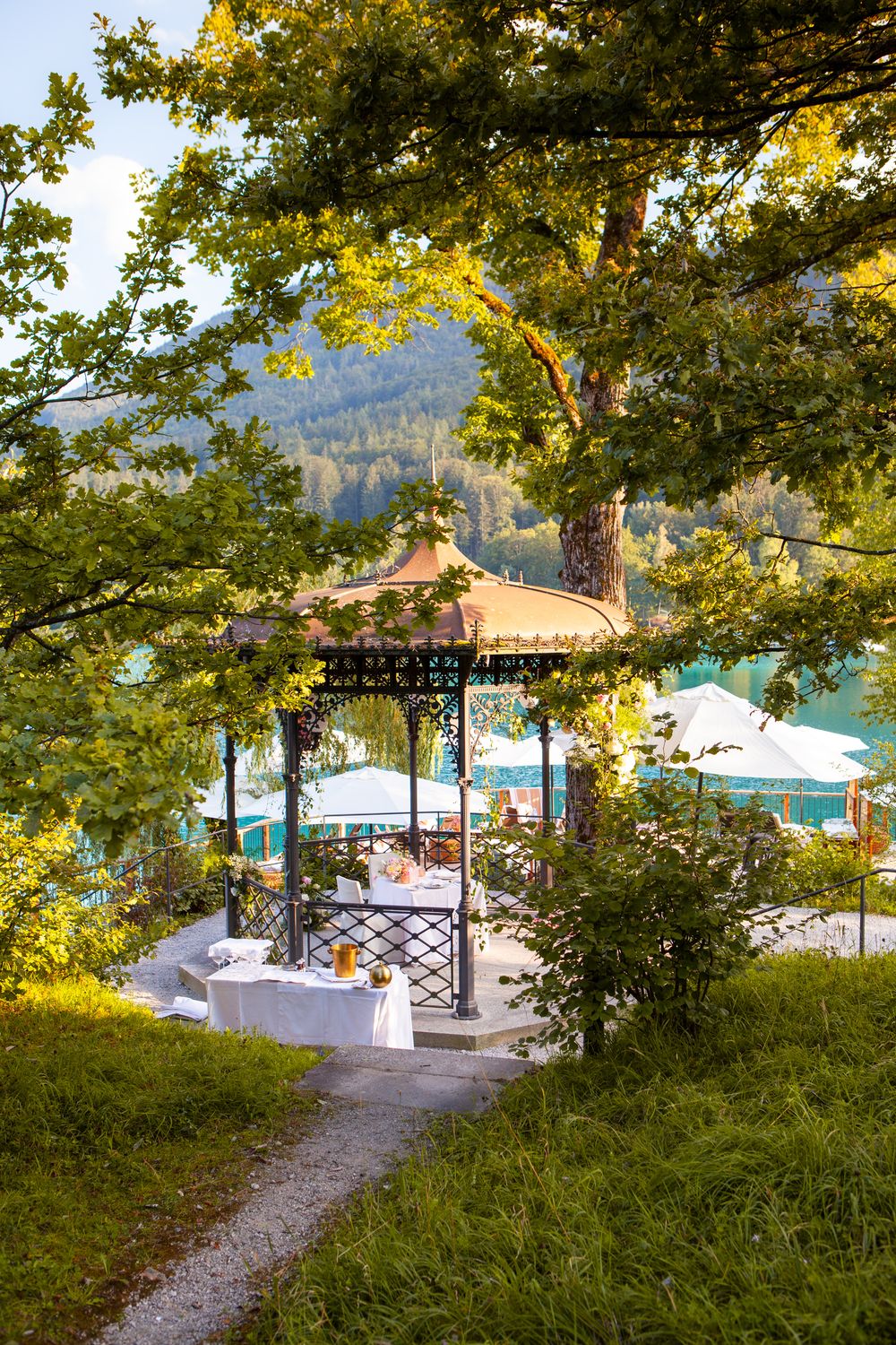 Elegant garden gazebo with ornate ironwork surrounded by lush trees overlooking a turquoise lake and mountain backdrop.