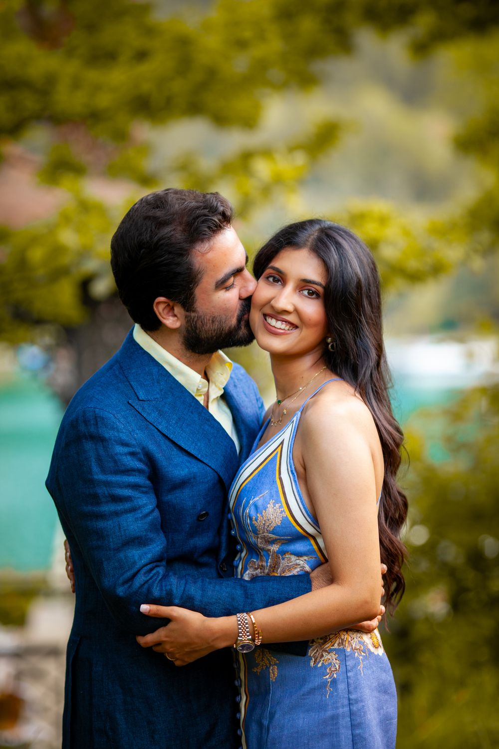 A man in a blue suit kisses a smiling woman in a blue embroidered dress outdoors.