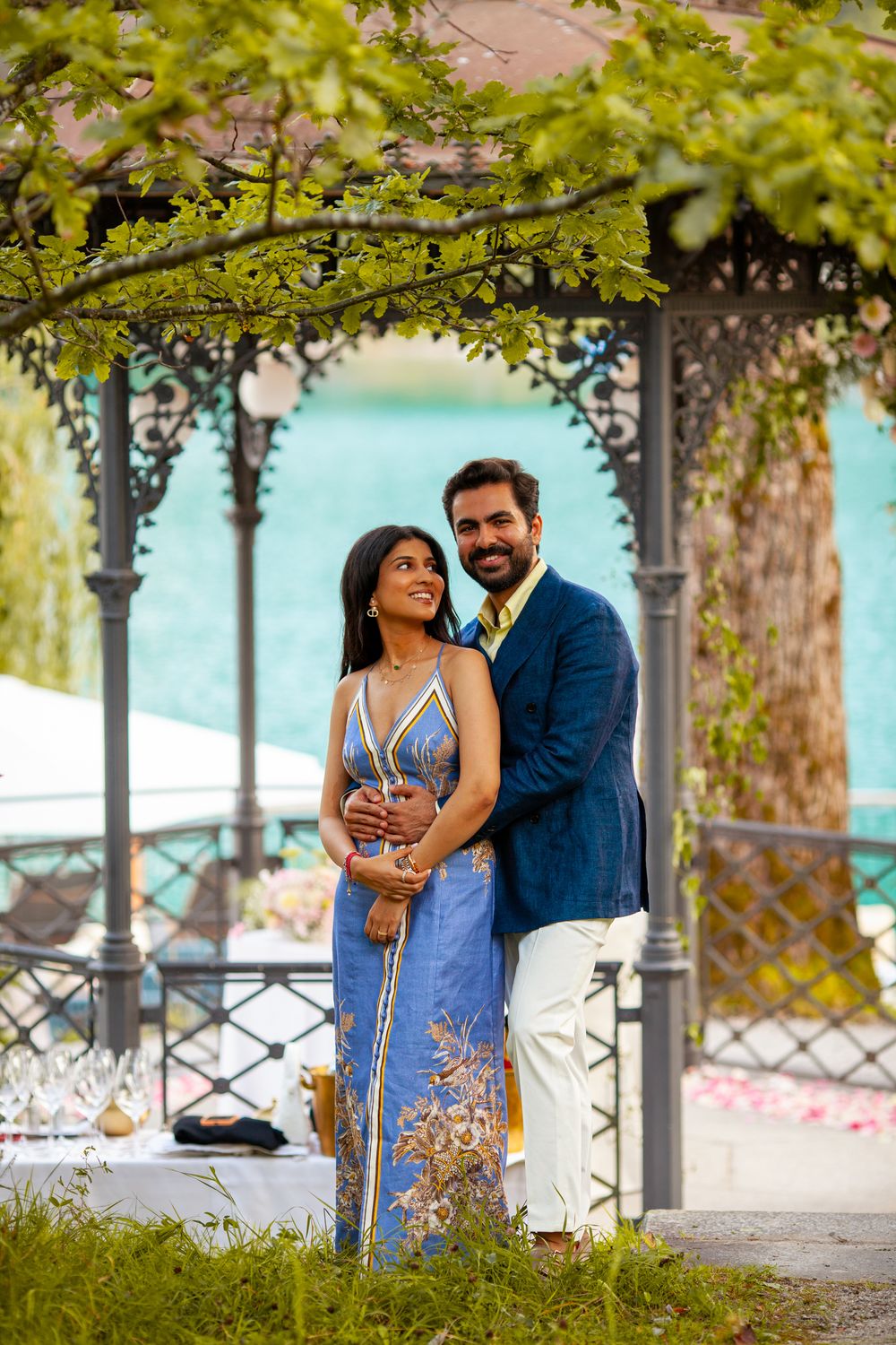 Couple in elegant attire embracing under ornate gazebo by turquoise lake, woman in blue floral gown, man in navy blazer.