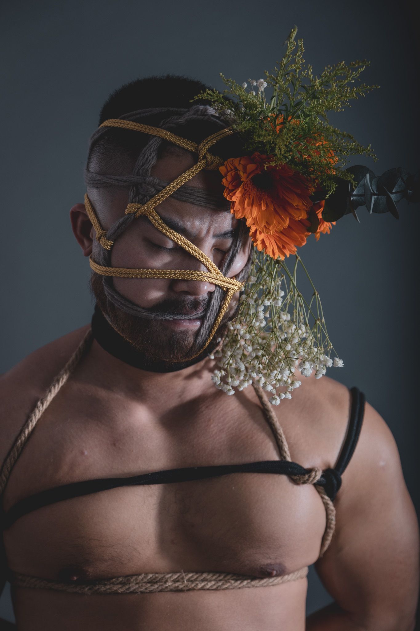 Bearded man with rope face bondage featuring orange marigold and baby's breath floral arrangement against gray background.