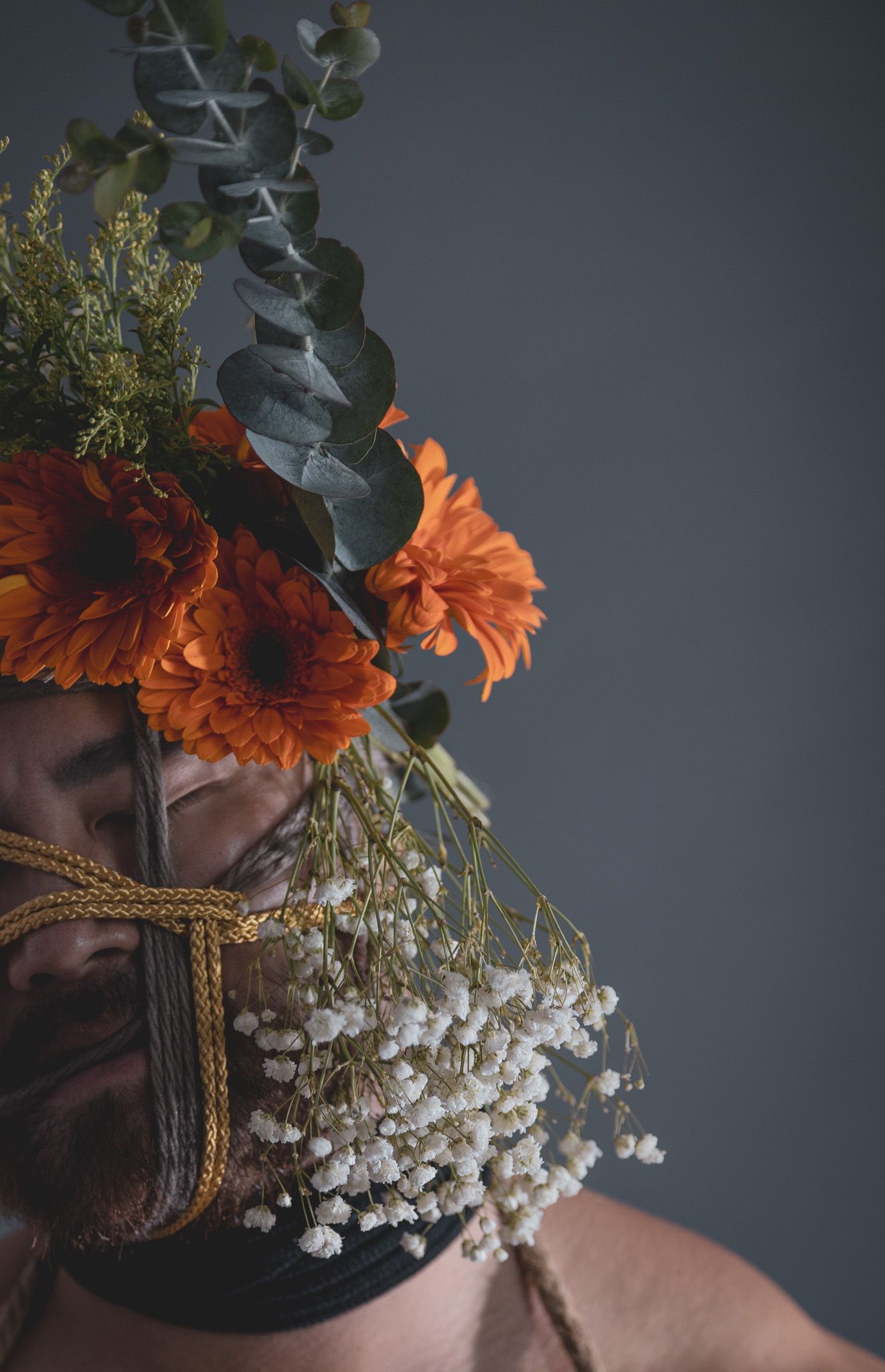 Close-up of person with rope face harness adorned with orange gerbera daisies, eucalyptus, and baby's breath flowers.