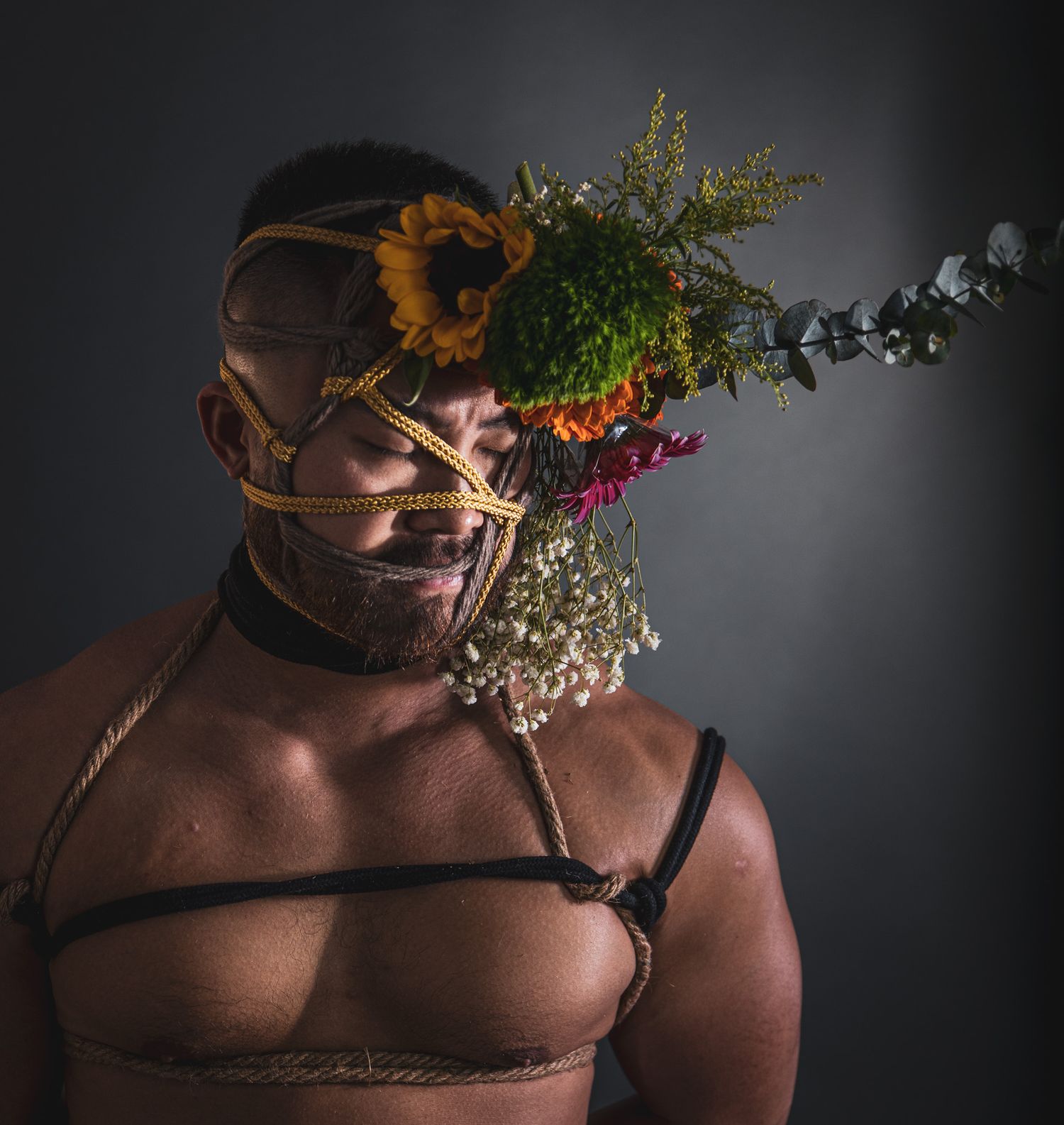 Studio portrait of a man with leather and rope face harness decorated with sunflowers and greenery.