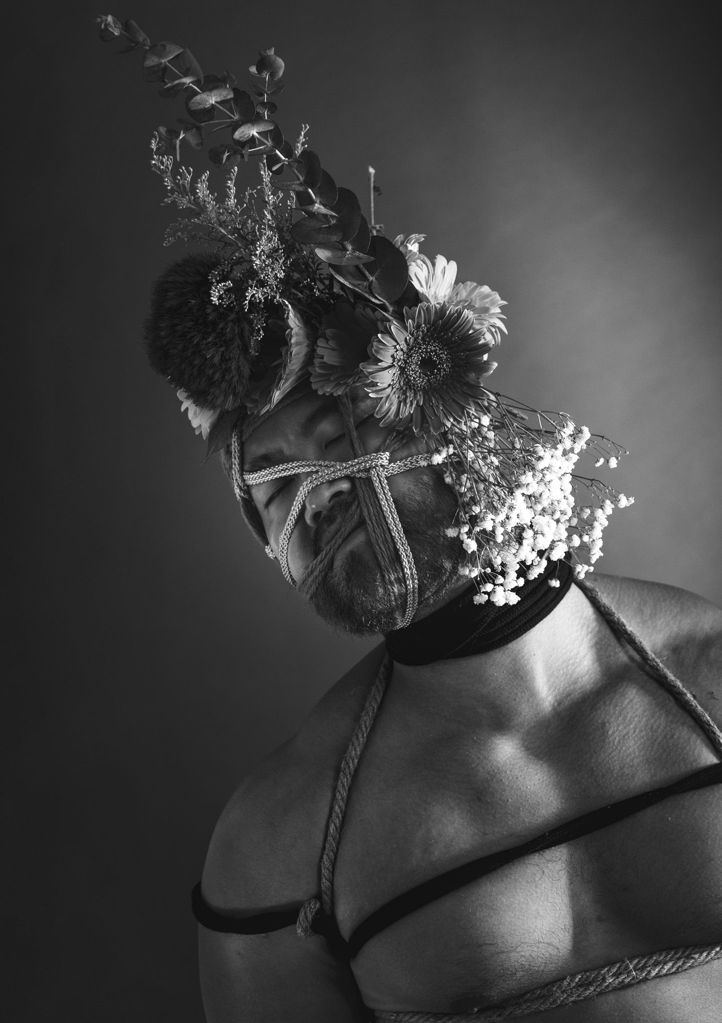 Black and white artistic portrait of a man with rope harness and floral crown covering his face.