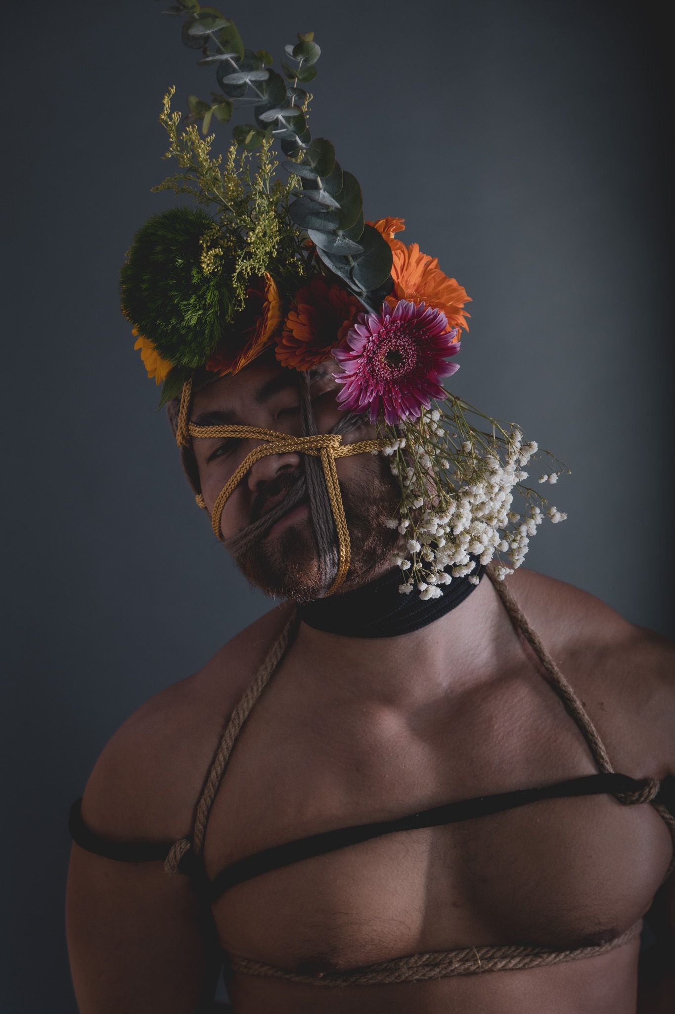 Studio portrait of a shirtless man with rope face binding decorated with colorful gerbera daisies.