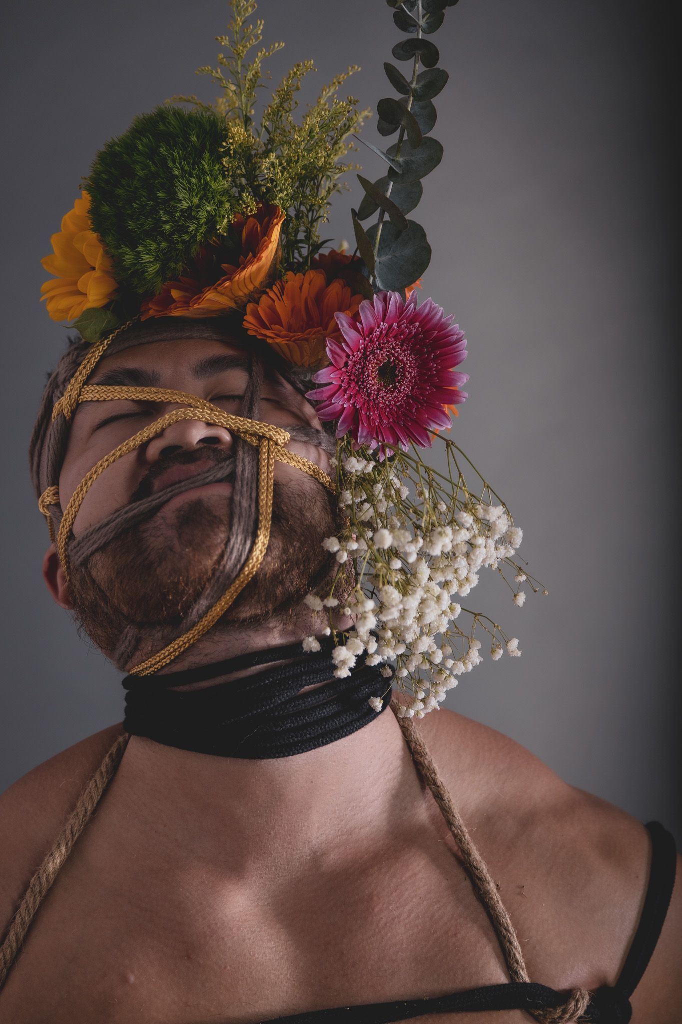 Bearded man with rope bondage on face adorned with colorful flowers including gerbera daisies and baby's breath.
