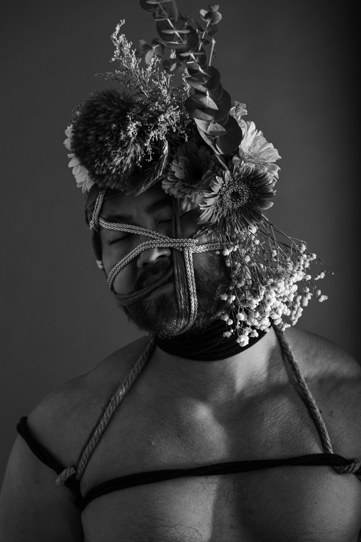 Black and white portrait of bearded man with rope face bondage decorated with sunflowers and wildflowers.