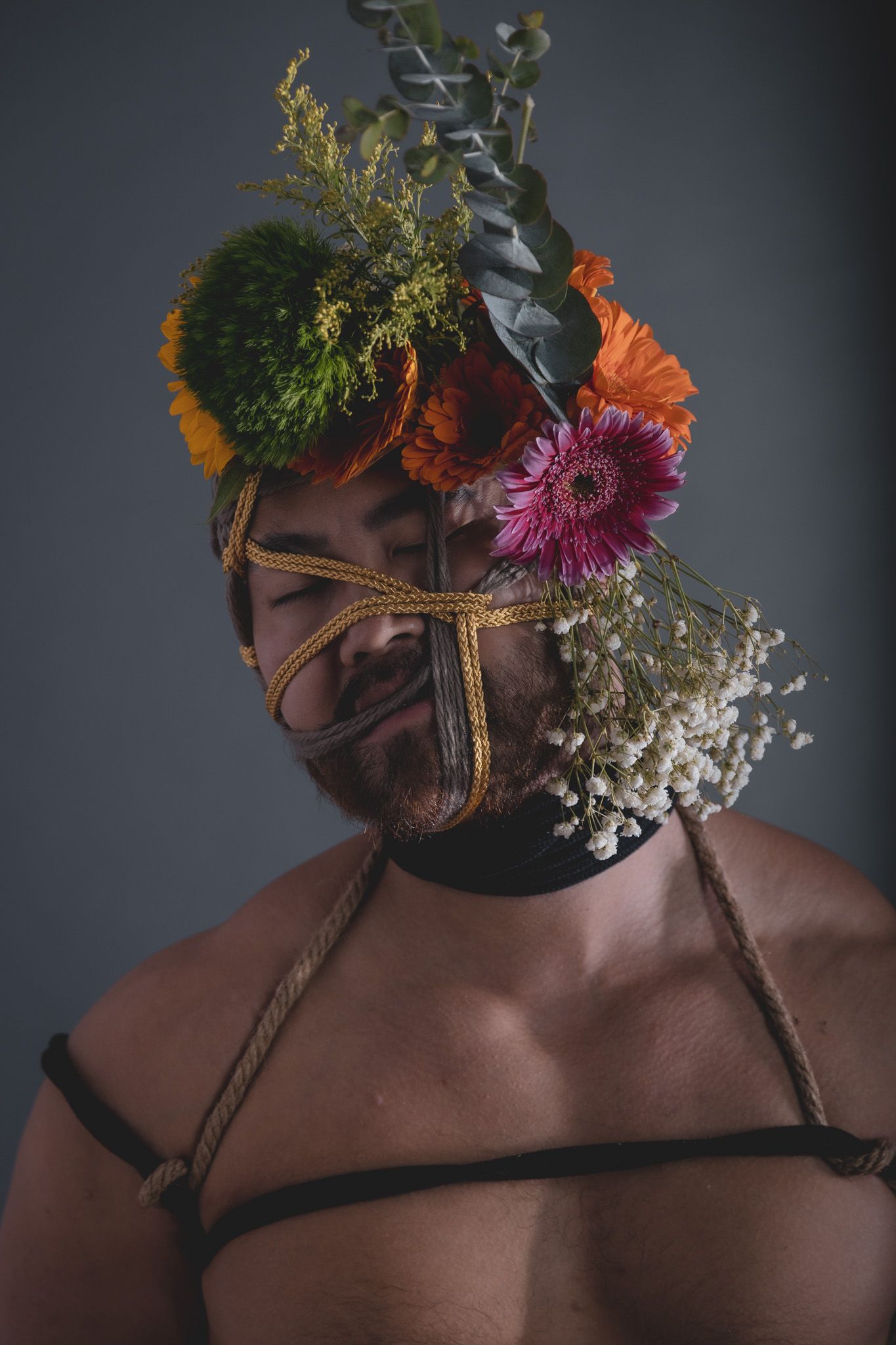 Smiling bearded man with rope face harness decorated with vibrant orange and pink gerbera daisies and greenery.