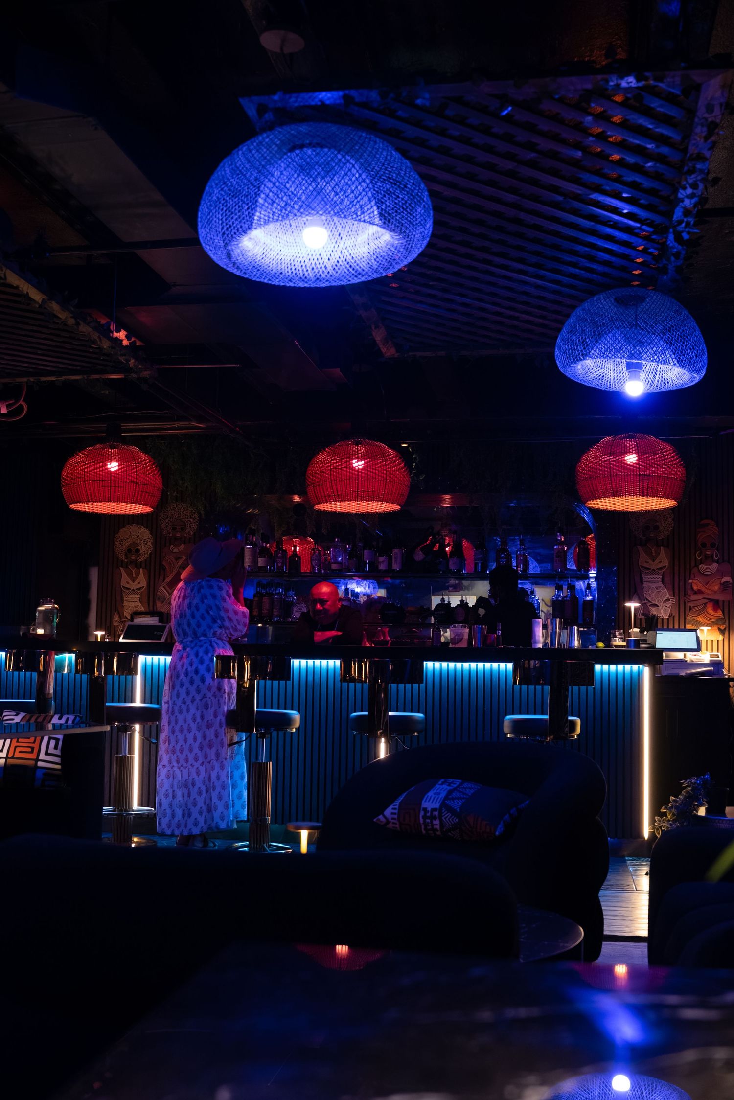 Dimly lit bar with blue and red wicker pendant lights, a bartender silhouette, and glowing blue counter accents.
