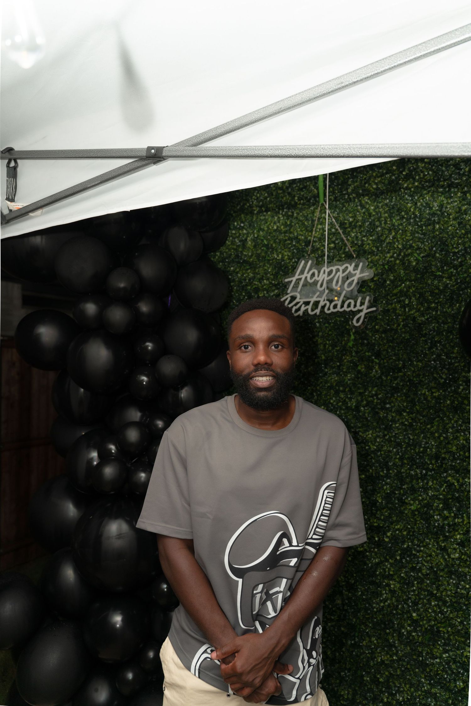 Person in gray t-shirt seated against green hedge wall backdrop with black balloons in event setting.