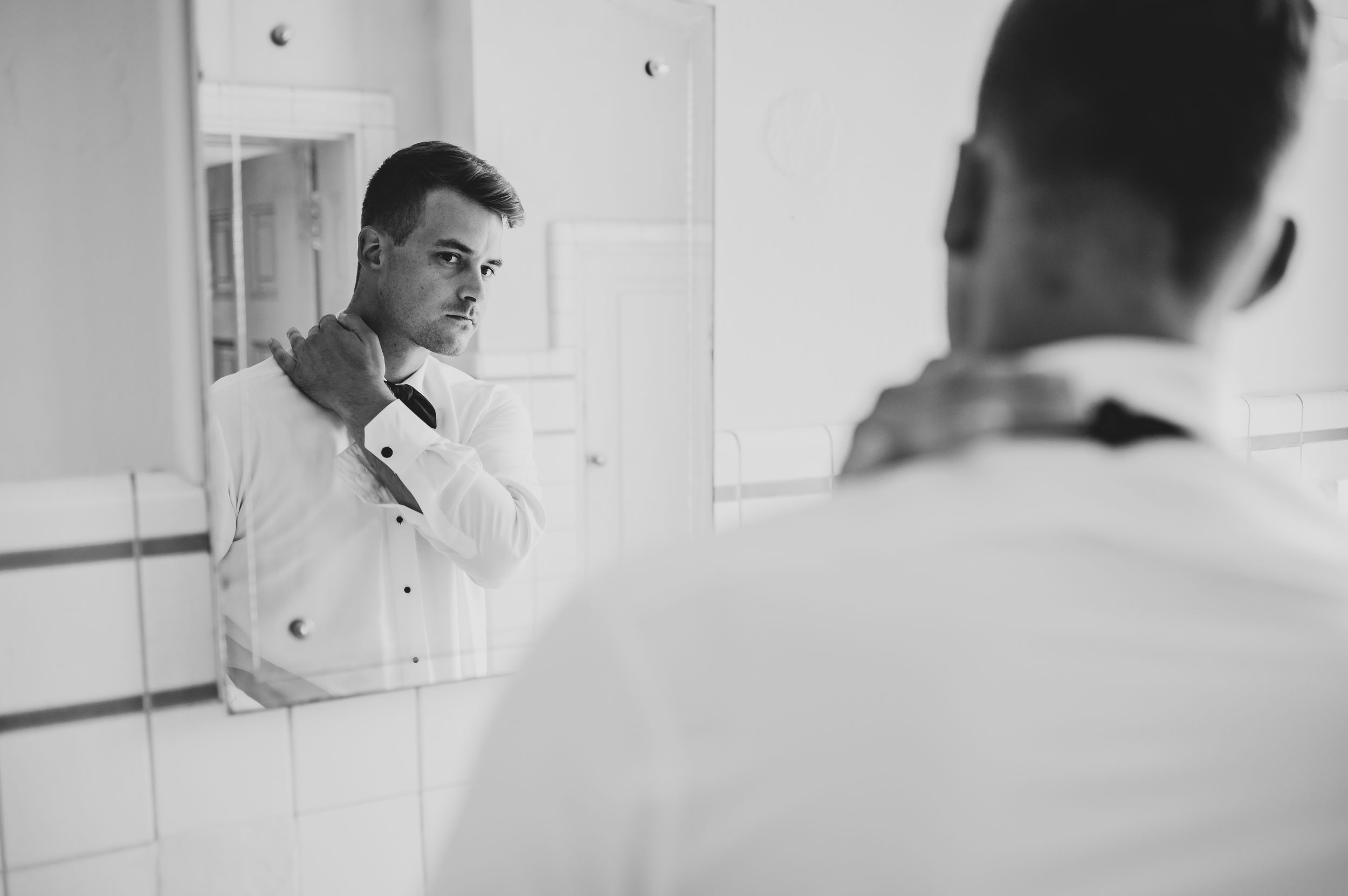Black and white photo of a man adjusting his tie in a bathroom mirror while getting dressed.