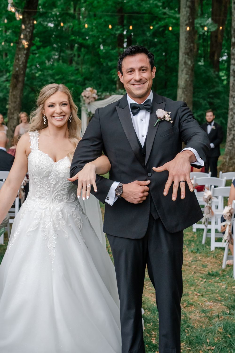 Bride in white lace gown and groom in black tuxedo showing wedding rings at outdoor forest ceremony.
