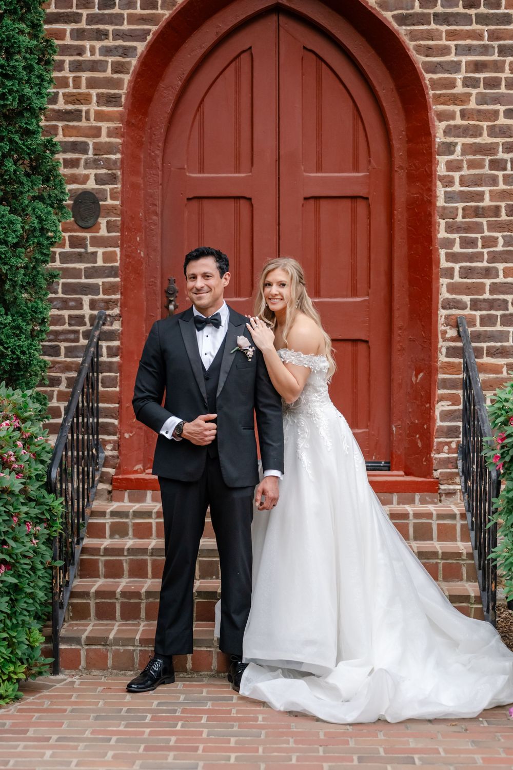 Bride and groom posing in front of a red arched church door on brick steps on their wedding day.