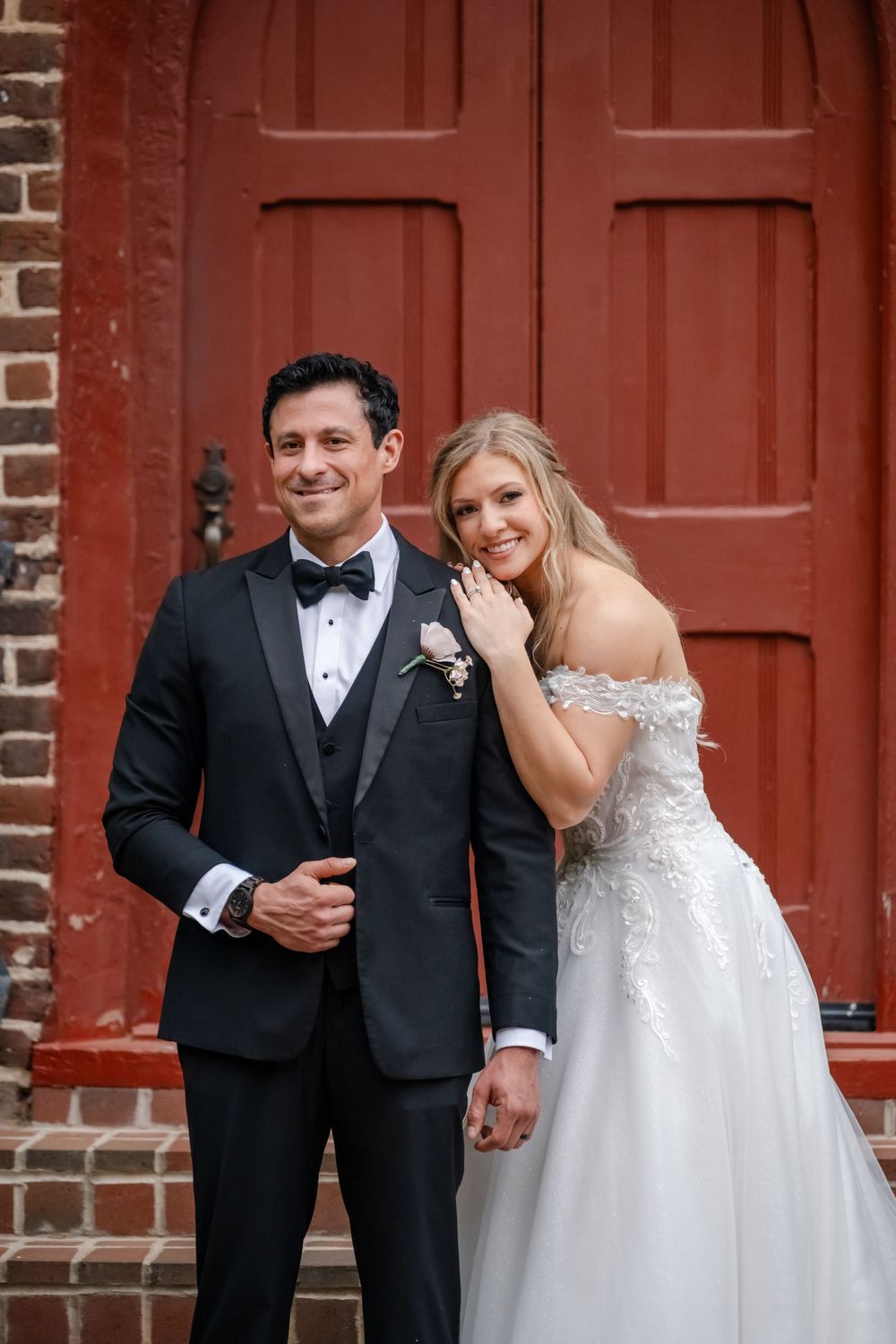 Bride and groom posing in front of a red door, she in a white gown and he in a black tuxedo.