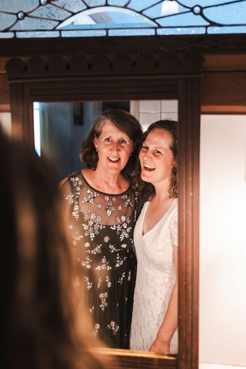 Two women smile joyfully in a wooden mirror reflection, one in a white wedding dress and one in a black floral gown.