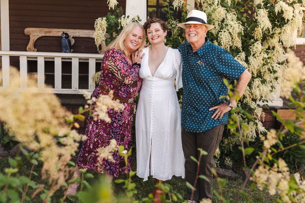Three women smiling together in a garden surrounded by white flowering bushes near a charming home.
