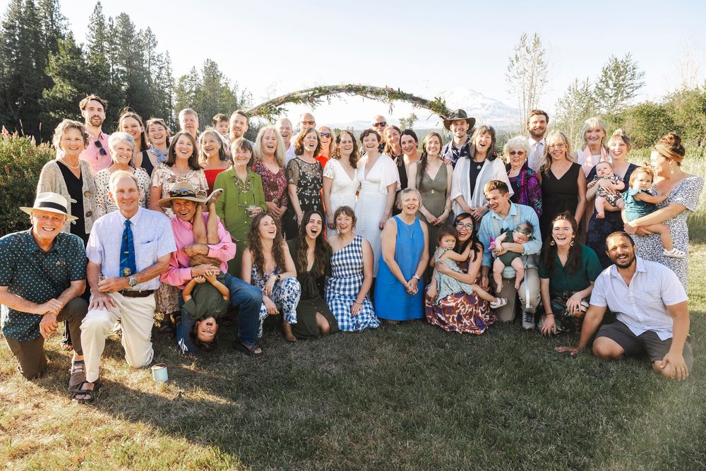 Large wedding party group photo outdoors under a floral arch with mountains in the background.