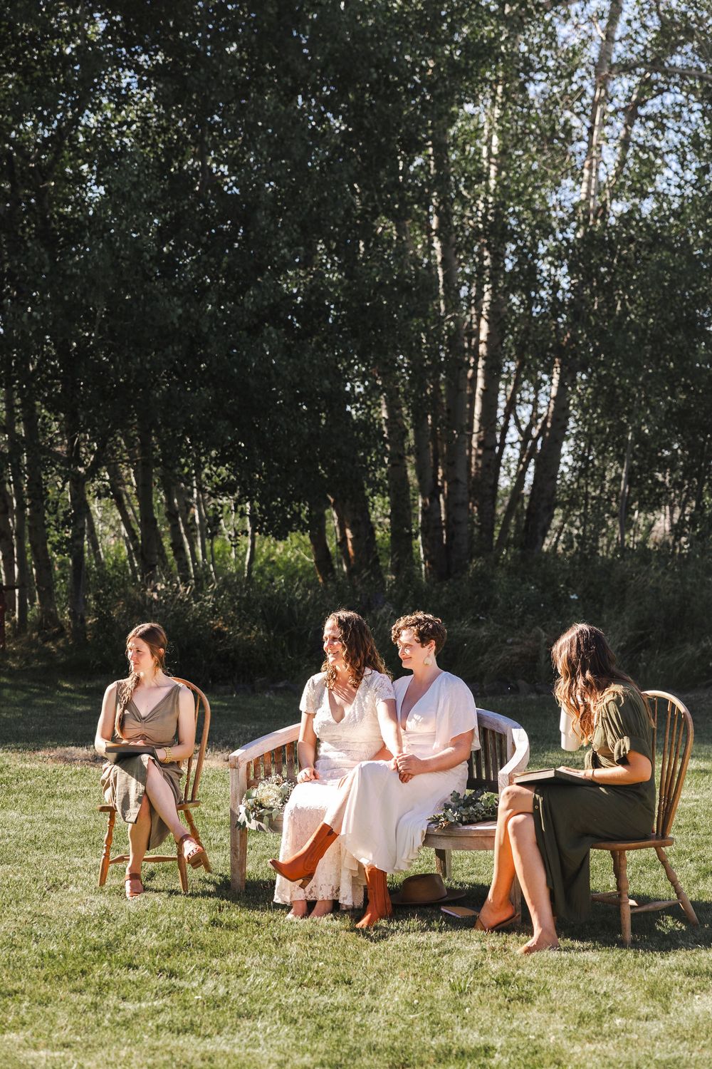 Four women sit outdoors on chairs and a bench surrounded by lush green trees during golden hour.
