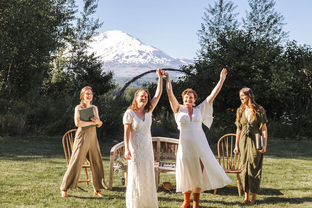 Two brides celebrate joyfully with raised hands surrounded by friends on a lawn with a scenic mountain view.