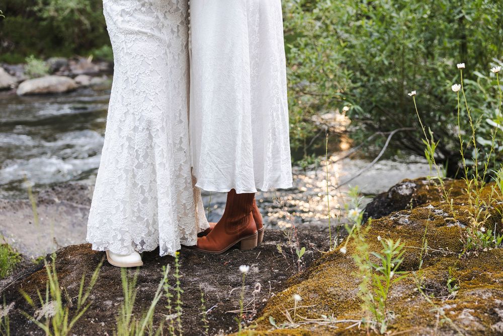 Close-up of bride in lace wedding dress and brown ankle boots standing on rocks near a flowing stream outdoors.