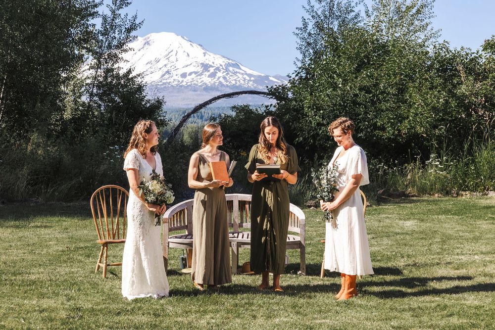 Four women in elegant attire stand on a lawn with a snow-capped mountain backdrop during an outdoor wedding ceremony.