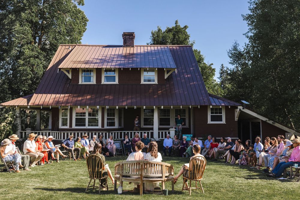 Guests gather outdoors on lawn chairs before a rustic red farmhouse with metal roof during a sunny outdoor event.