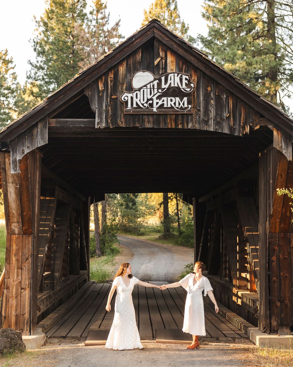 Two brides in white dresses hold hands under a rustic covered bridge at Trout Lake Farm.