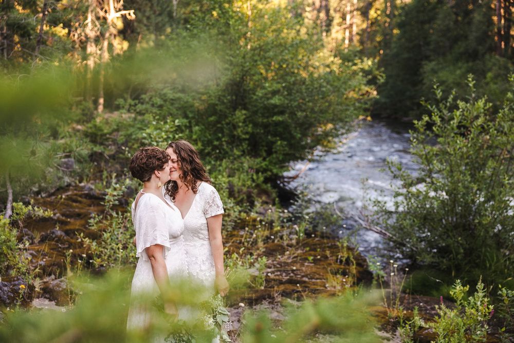 Two brides in white dresses share an intimate moment beside a forest stream in lush greenery.