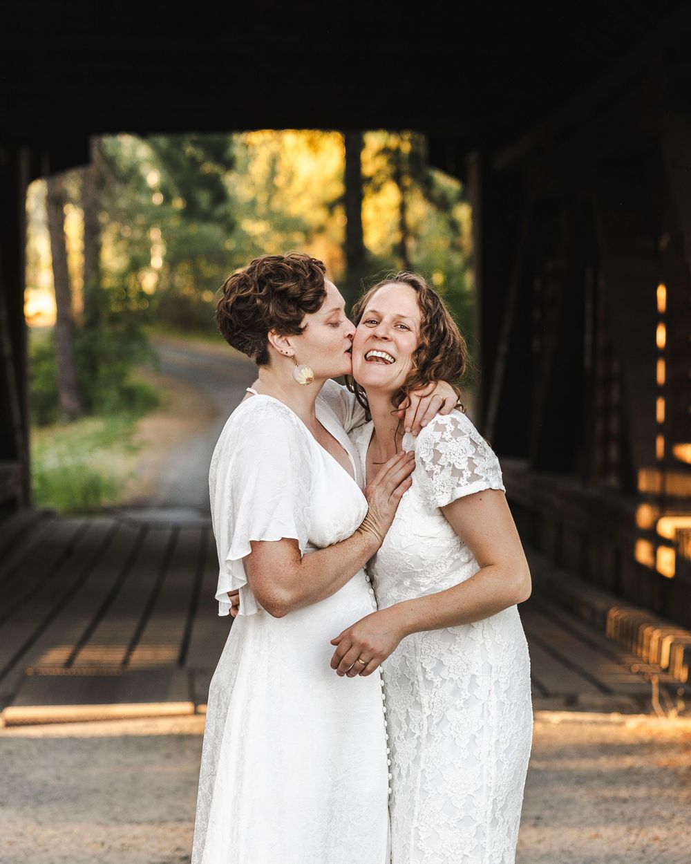 Two brides share a joyful kiss under a rustic covered wooden bridge in golden forest light.