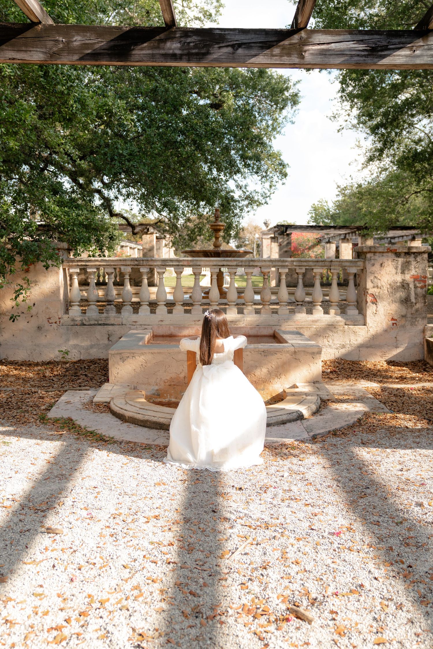 Young girl in white dress stands before ornate stone balustrade and fountain in sunlit garden.