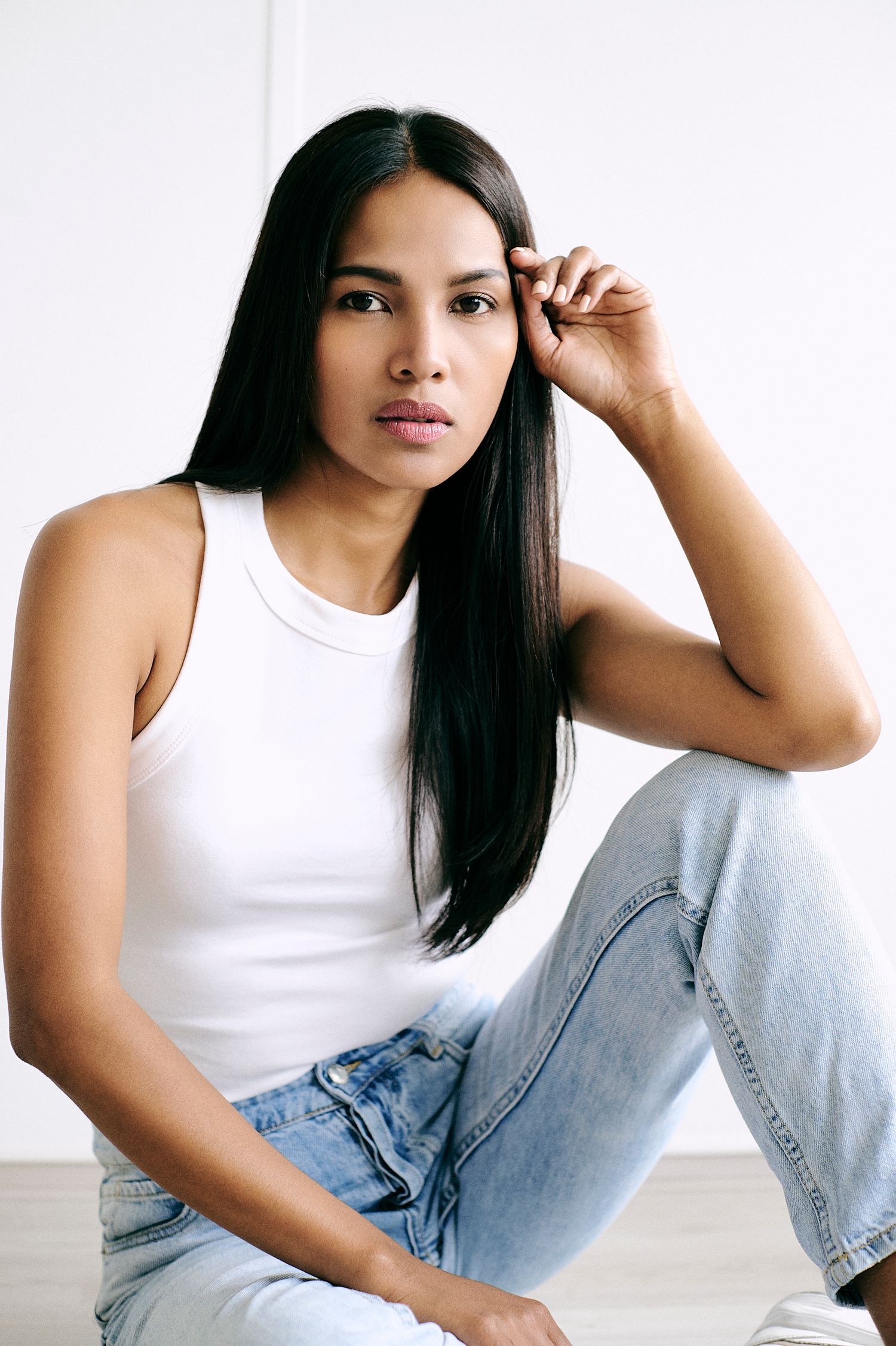 Young woman with long straight black hair resting chin on hand, wearing white top against white background.