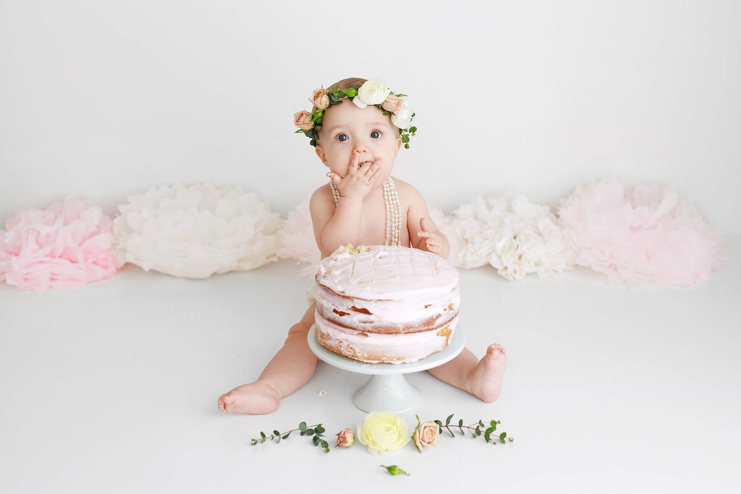 Baby girl in flower crown doing a cake smash with a pink cake on white backdrop.