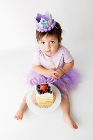Baby girl in purple tutu and crown sitting with a fruit-topped first birthday cake.